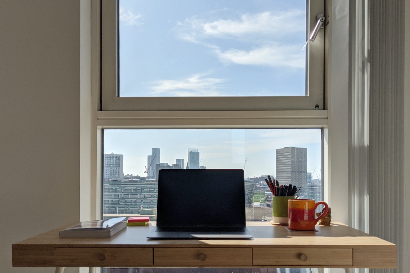 Laptop open on a desk in front of a window, out that window a blue sky and Manchester skyline